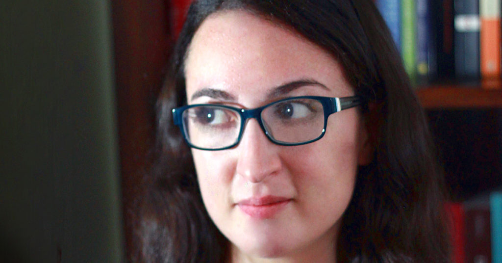 Dark haired woman with teal framed glasses looks to the left.