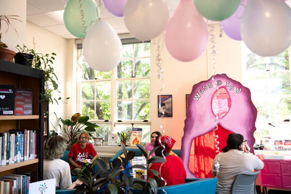 A group of people converse in a library with balloons hanging from the ceiling, surrounded by books and plants.