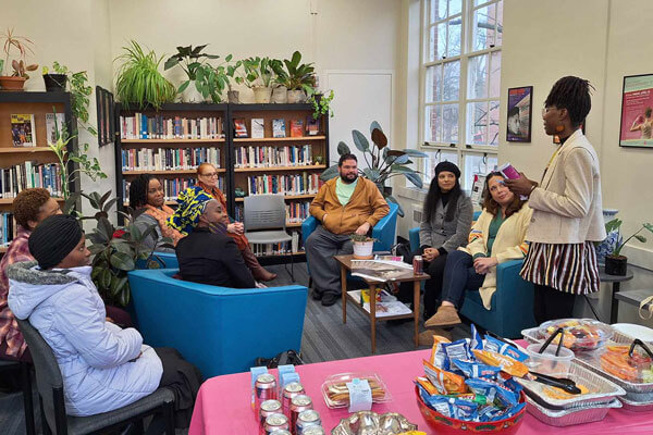 A group of people in a library setting seated in a semi-circle, with one person standing and speaking. A table with snacks is in the foreground.