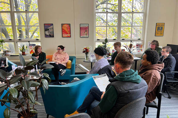 A group of people having a meeting in a well-lit room with large windows and colorful posters on the walls.