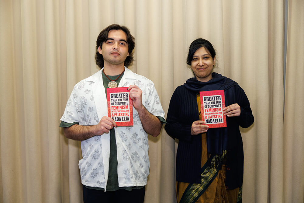 Two people holding a book against a beige curtain backdrop.