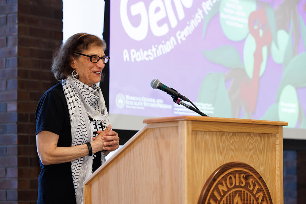 Person speaking at a podium with a purple presentation slide in the background.
