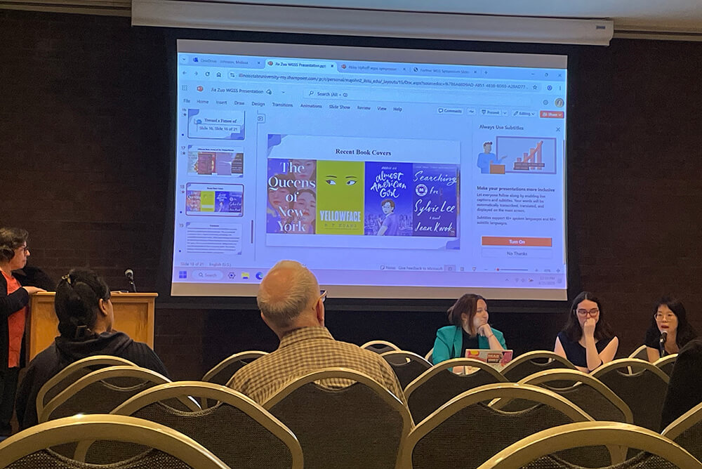 Lecture room with a PowerPoint presentation on book covers displayed on a large screen. Four presenters are at the front.