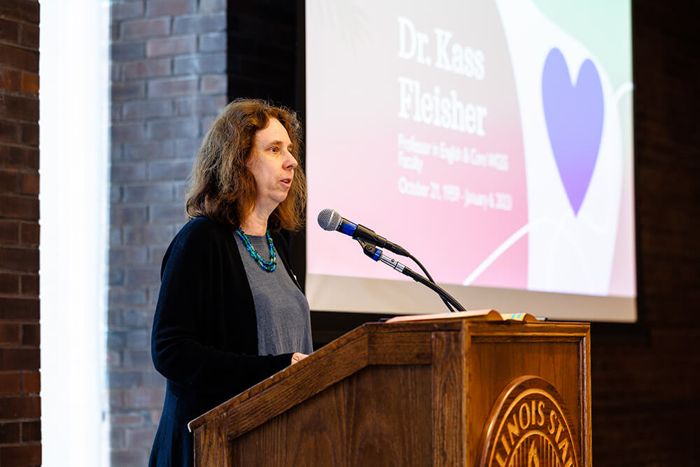 A person speaks at a podium with a screen displaying text and a heart graphic behind them.