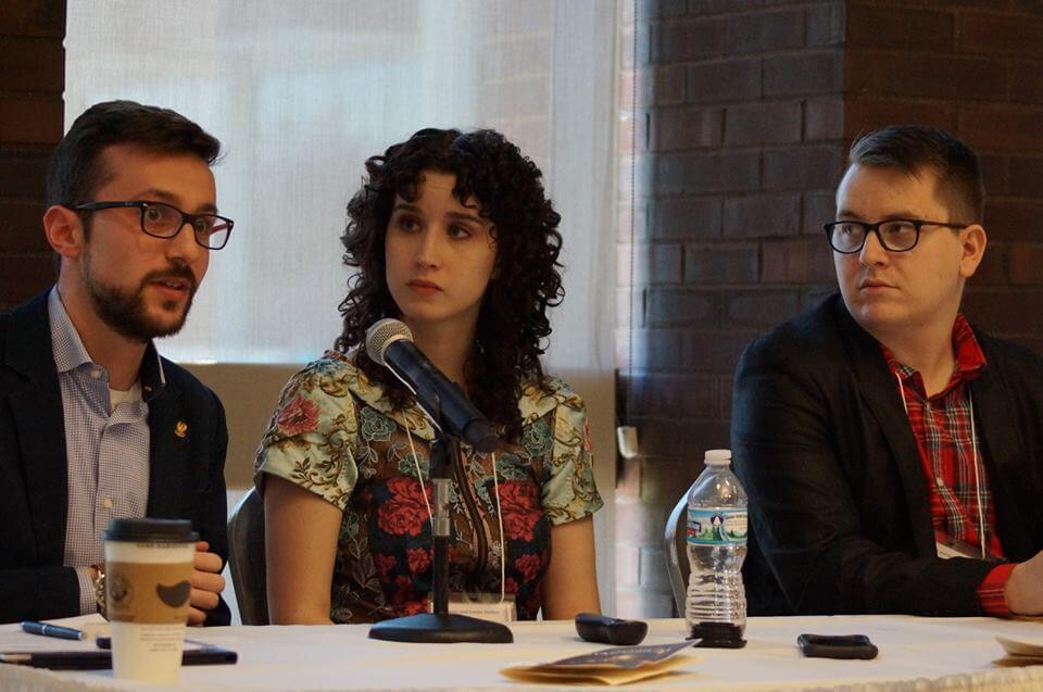 Three people seated at a panel discussion table, with microphones and drinks in front of them.